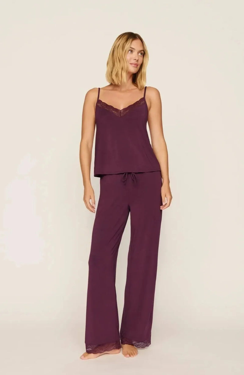 A woman modeling a two-piece pajama set consisting of a spaghetti-strap camisole top and matching wide-leg pants, both in a deep plum purple with a small white floral pattern, against a plain off-white background.